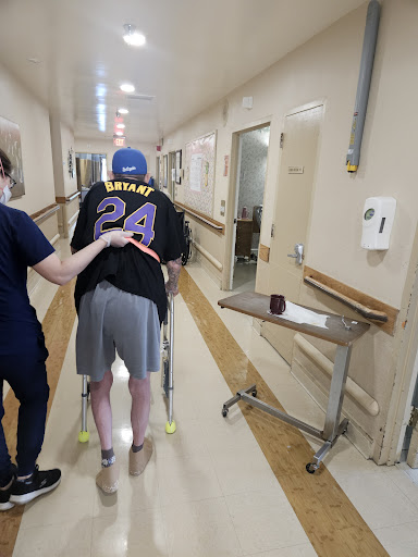 A caregiver assists a patient walking with a walker in a hospital corridor.