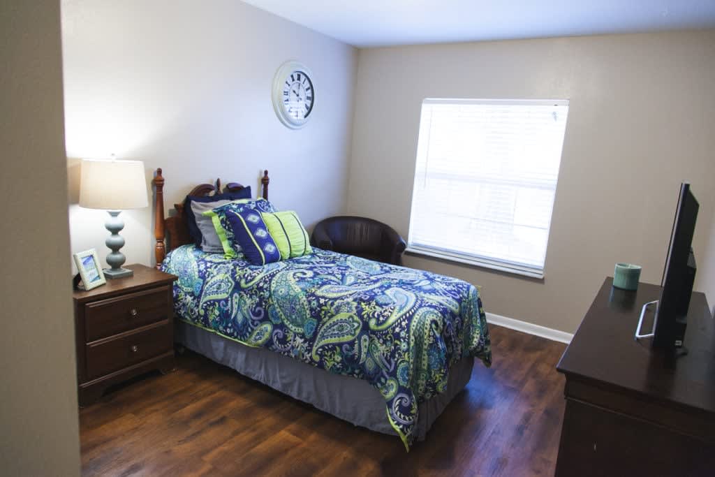 A cozy bedroom with a single bed and colorful bedding, featuring a lamp, bedside table, and window.
