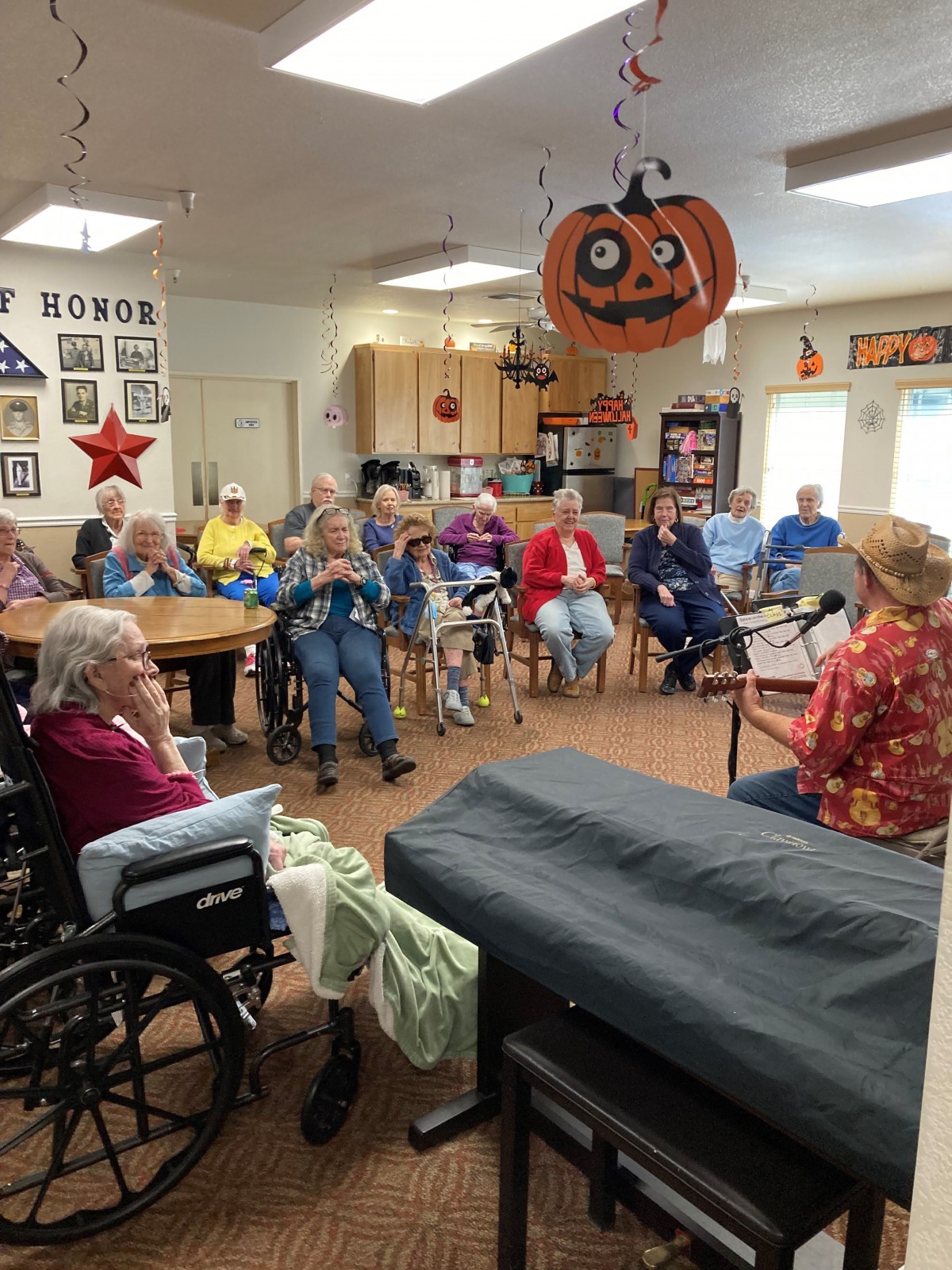 A group of elderly people gathered indoors for a community event, with Halloween decorations hanging from the ceiling.