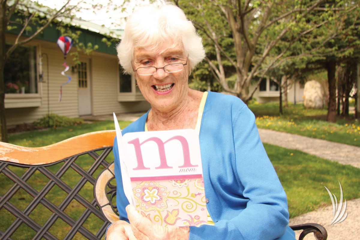 An elderly woman sitting outdoors, smiling and holding a book.