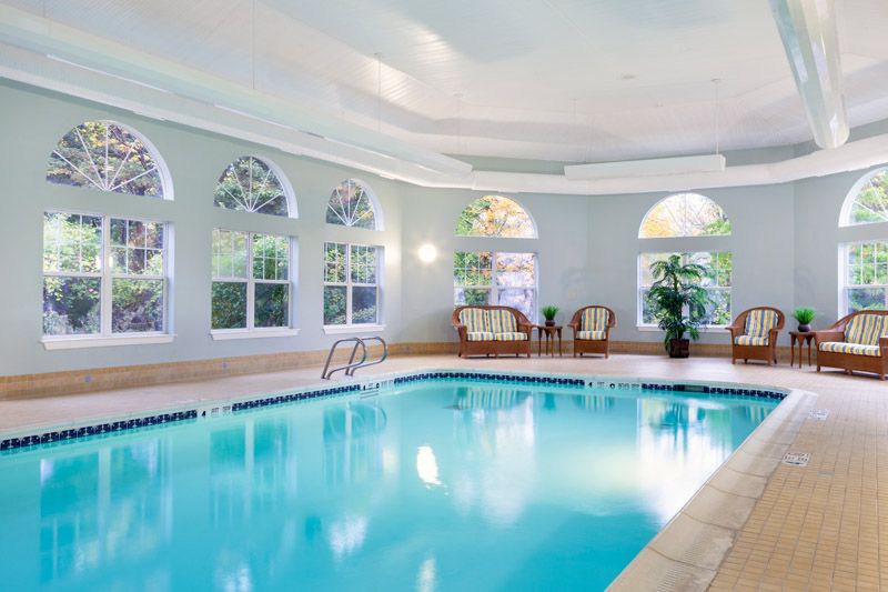 A serene indoor swimming pool area with bright lighting, surrounded by arched windows, chairs, and potted plants.