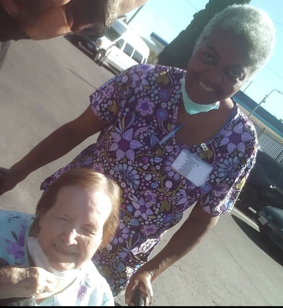 A smiling caregiver stands beside an elderly person in a wheelchair on a sunny day.
