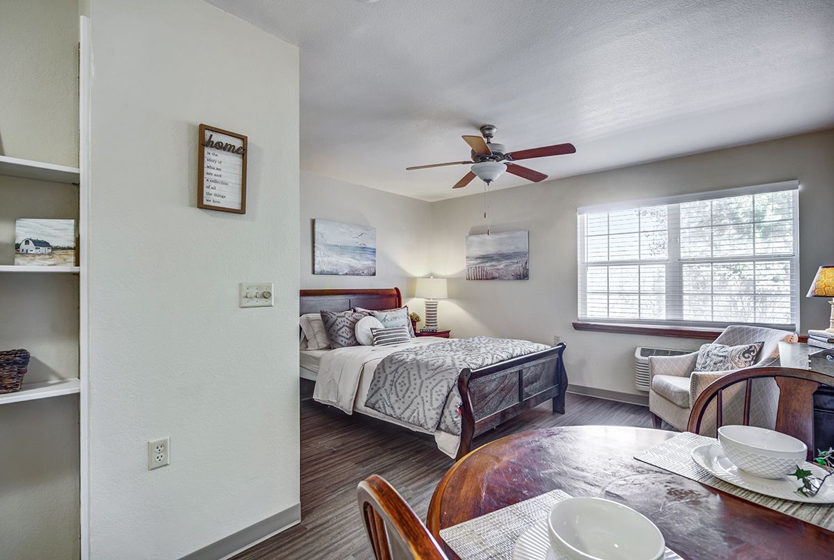 A cozy bedroom with a bed, table, ceiling fan, and artwork on the walls.