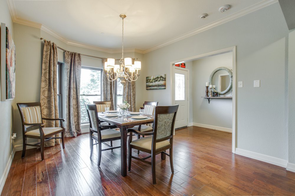 A cozy dining room with a wooden table and chairs, illuminated by a chandelier, featuring hardwood floors and elegant curtains.