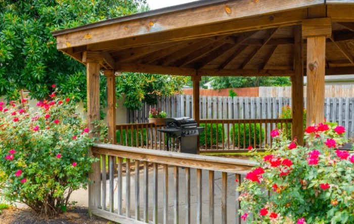 A wooden gazebo in a garden with vibrant roses and a grill.