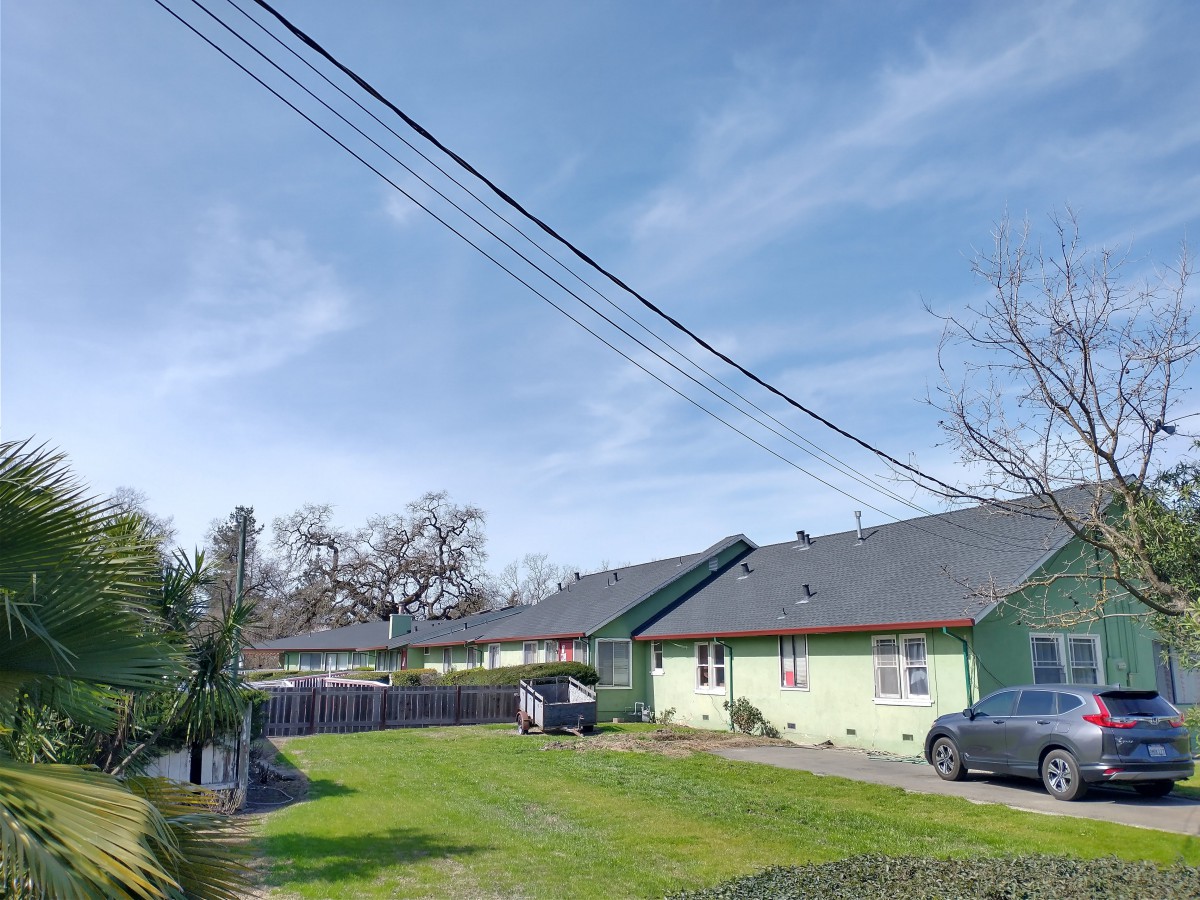 A suburban residential area with houses, a green yard, and parked cars under a blue sky.