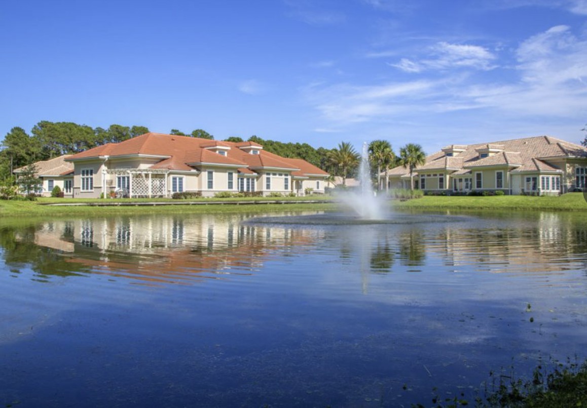 A peaceful residential area with houses surrounding a lake with a fountain under a clear blue sky.