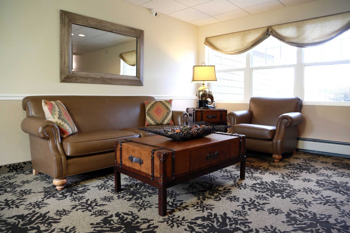 A cozy living room with a leather sofa, armchair, and a wooden coffee table adorned with decor. A large window allows natural light to fill the space, complemented by a warm lamp on the side.