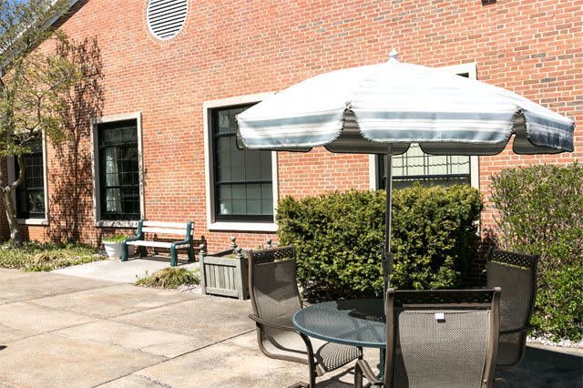 A sunny patio area with tables and chairs, featuring a large white umbrella and a backdrop of a brick building.