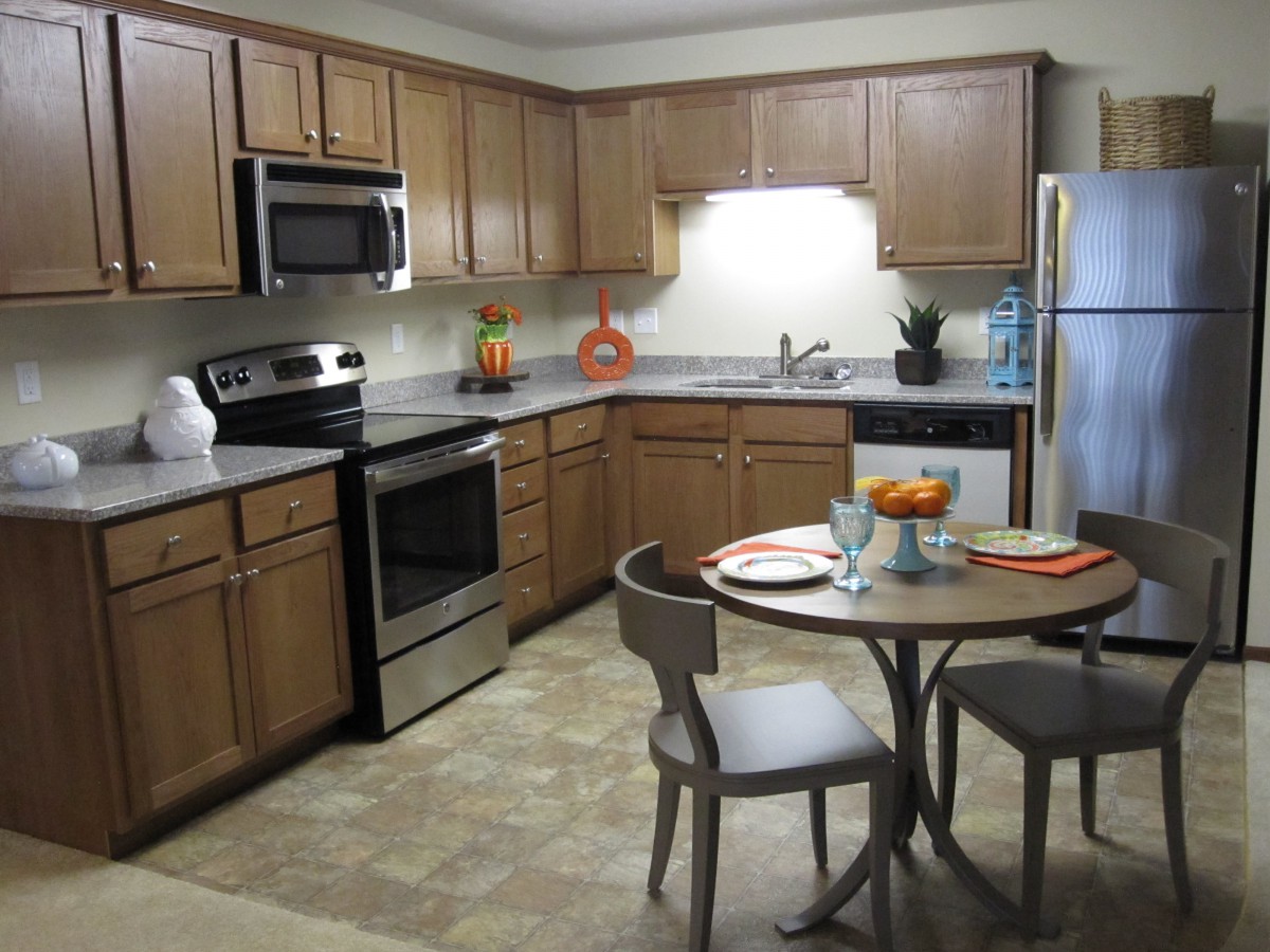 A modern kitchen with wood cabinets and stainless steel appliances, featuring a small dining area with a table and chairs.