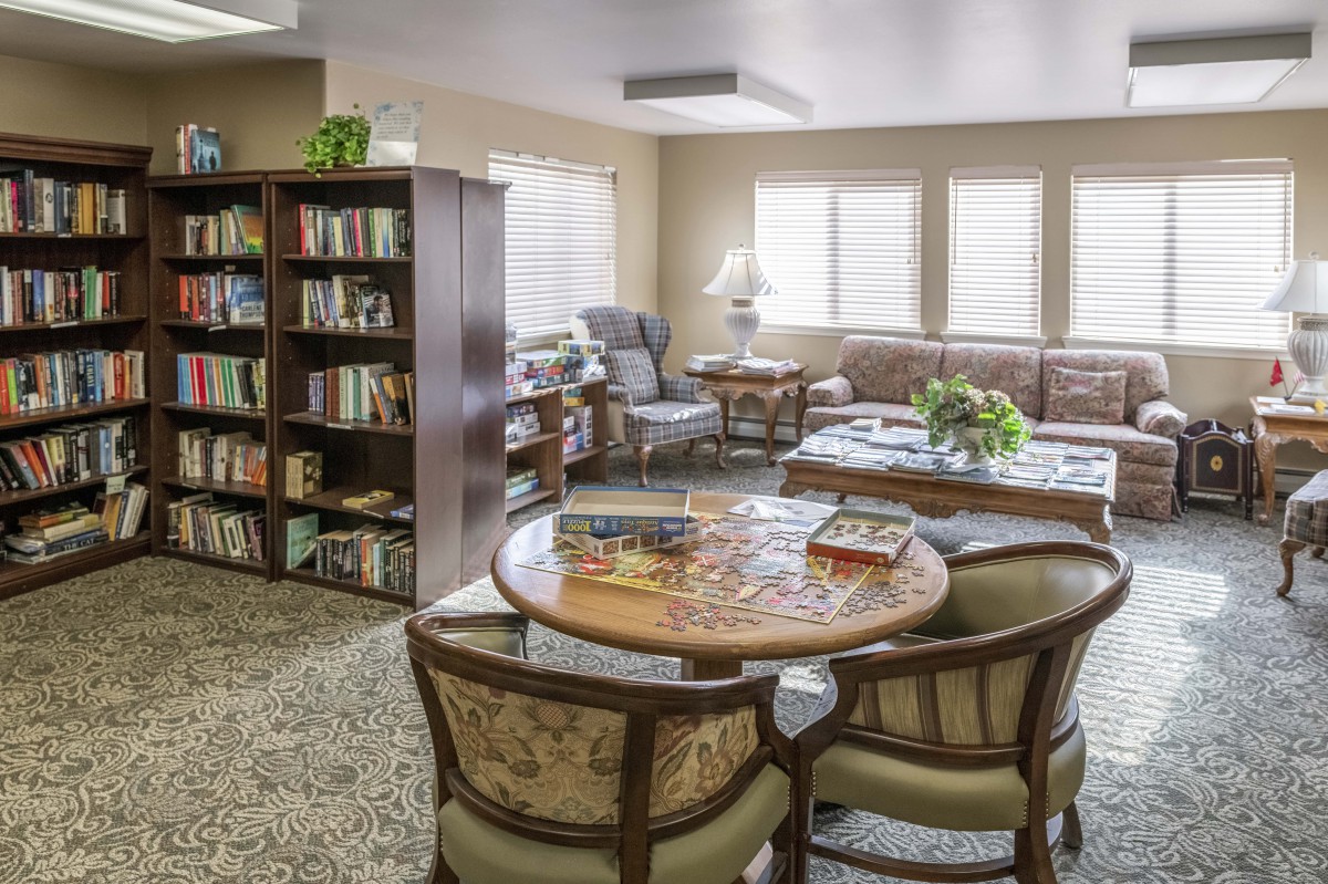 A cozy library room with bookshelves, a seating area, and a round table surrounded by chairs.