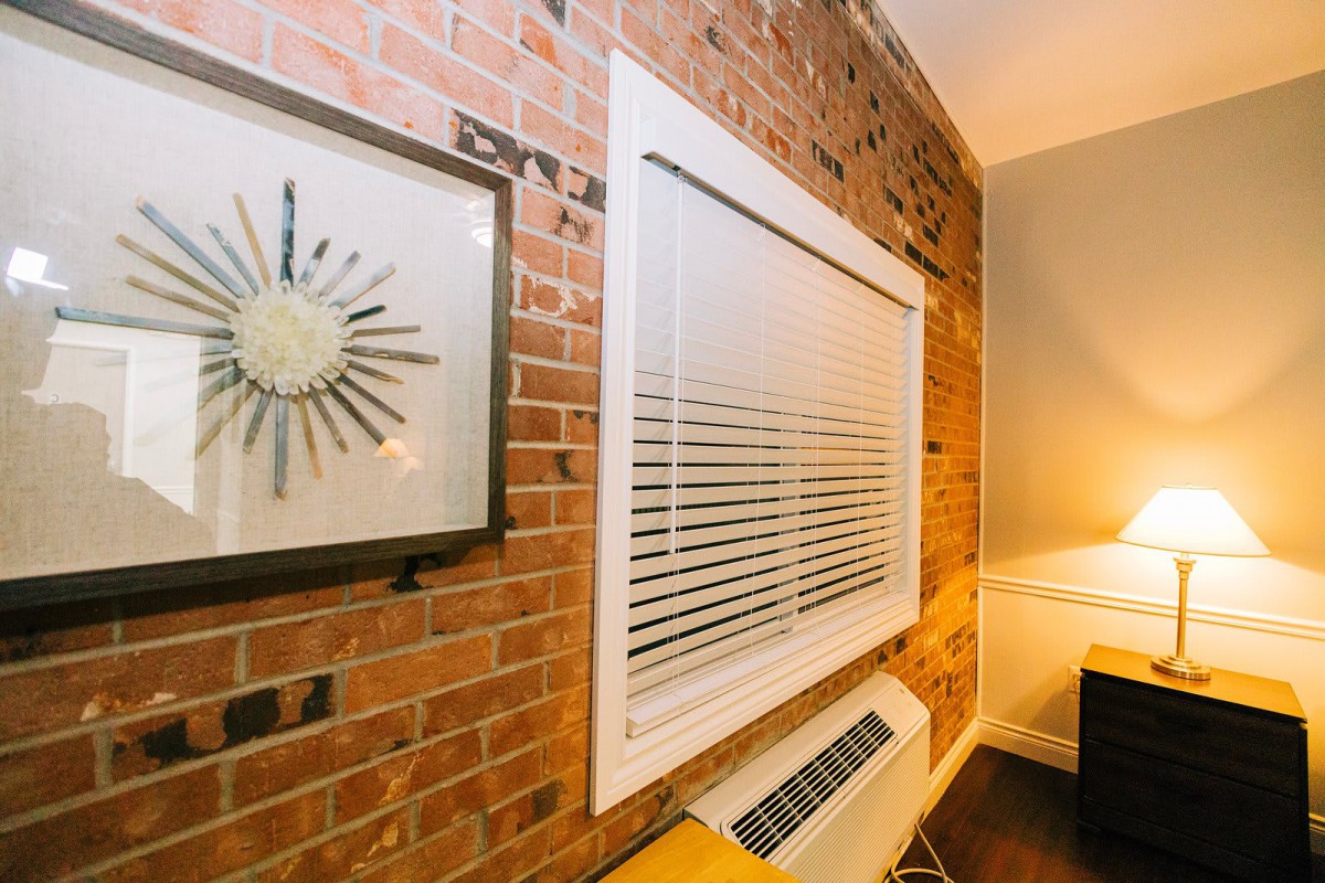 A cozy interior space featuring a brick wall, window with blinds, and a stylish clock above a modern air conditioner unit.