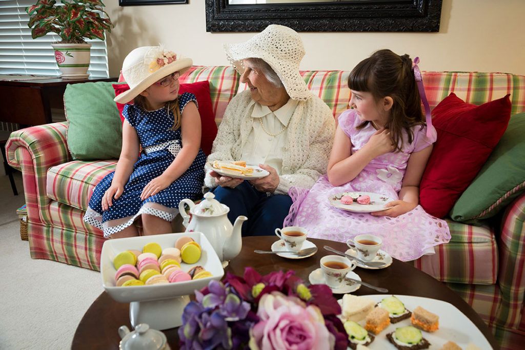 An elderly woman and two children enjoy a tea party with cakes in a cozy living room setting.