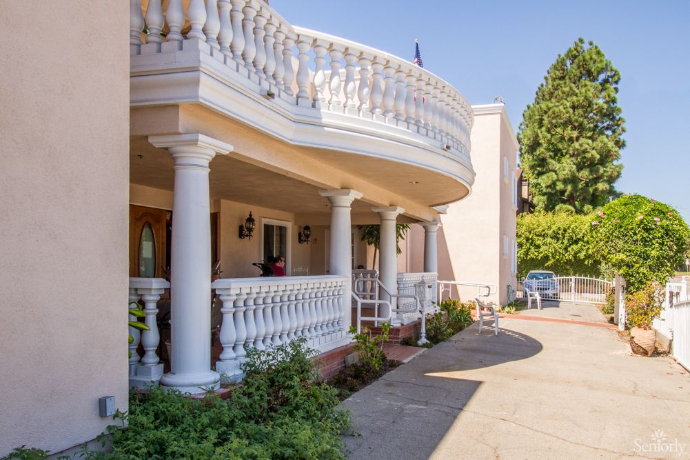 A residential building with a grand porch featuring columns and balustrades, alongside a driveway and surrounded by greenery.