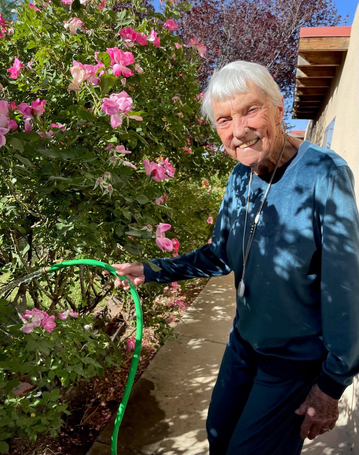 An elderly woman happily waters flowers with a garden hose on a sunny day.
