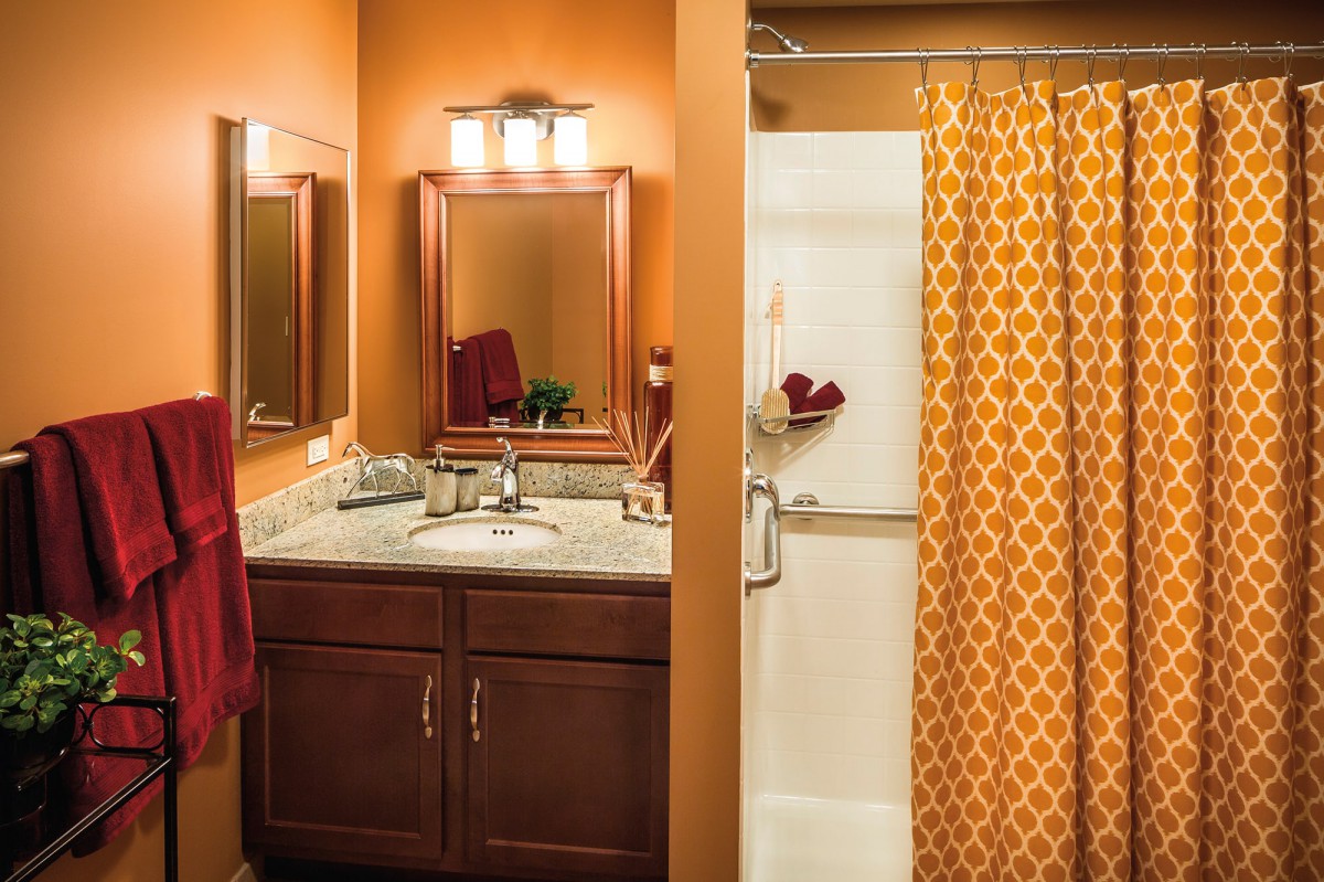 A warmly decorated bathroom with orange walls, featuring a sink, mirror, and a shower with a patterned curtain.