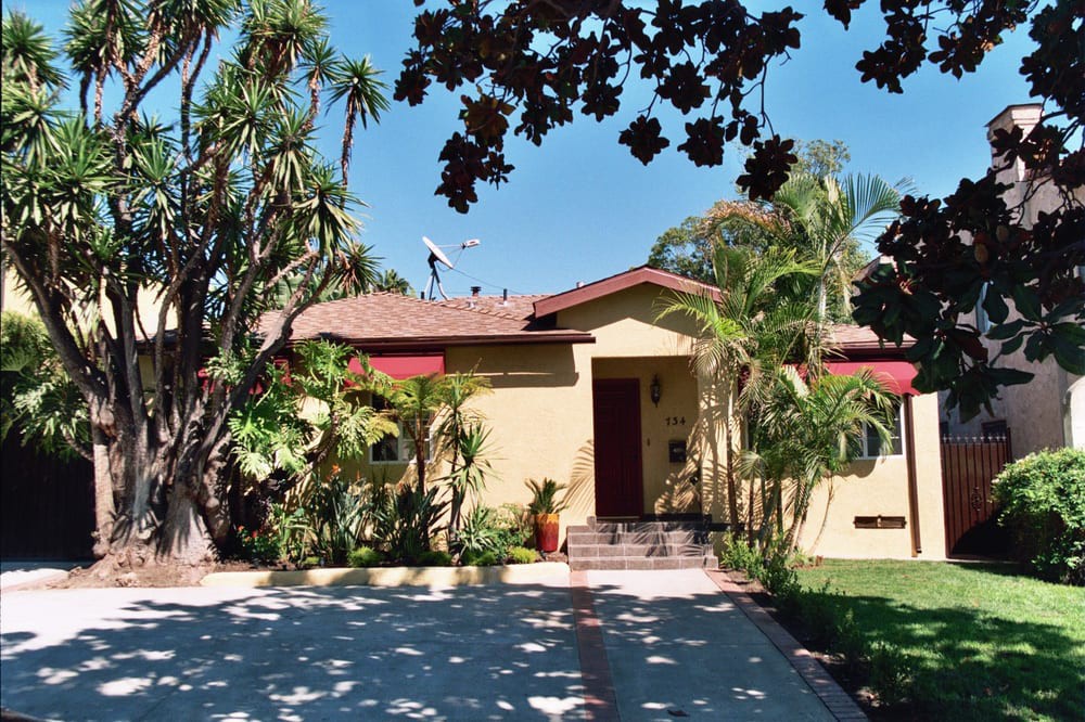A suburban house surrounded by tropical plants and trees, with a driveway in front on a sunny day.