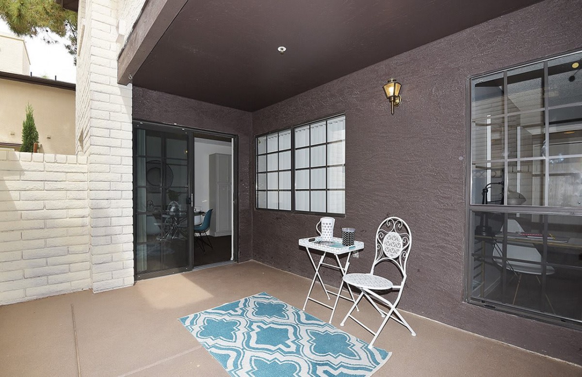 A cozy patio features a small table with two chairs, a decorative rug, and is enclosed by large windows and a sliding glass door.