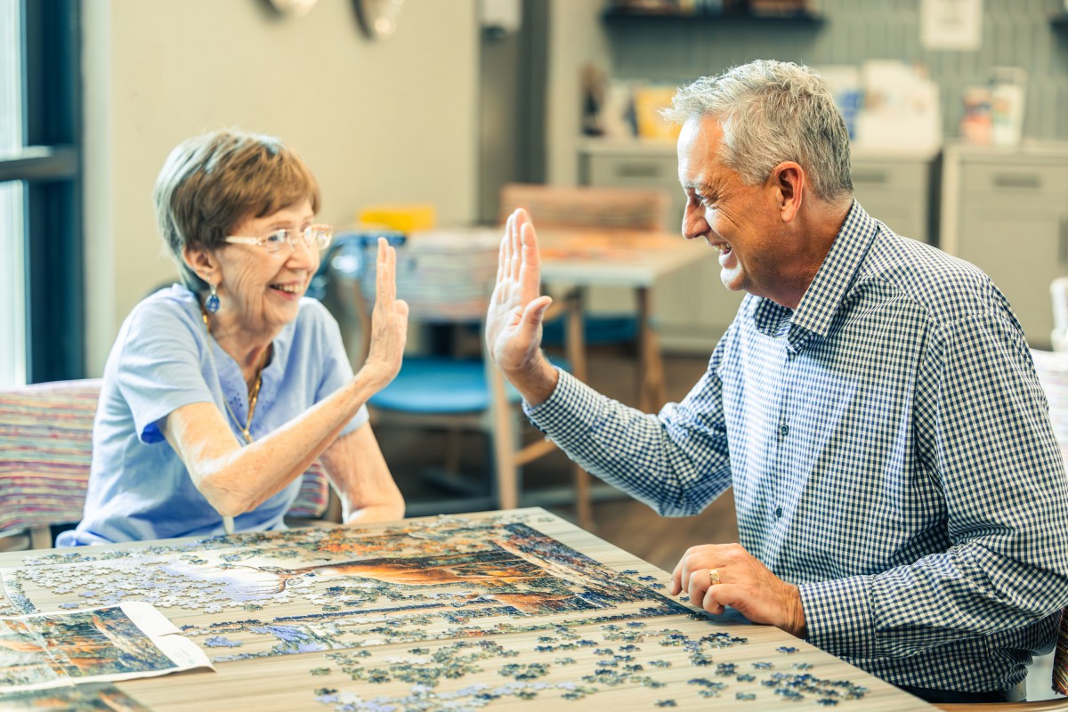An elderly couple shares a joyful high-five while working on a puzzle together indoors.