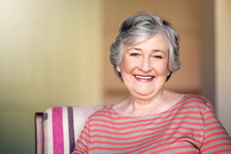 A smiling elderly woman sitting indoors wearing a striped shirt.