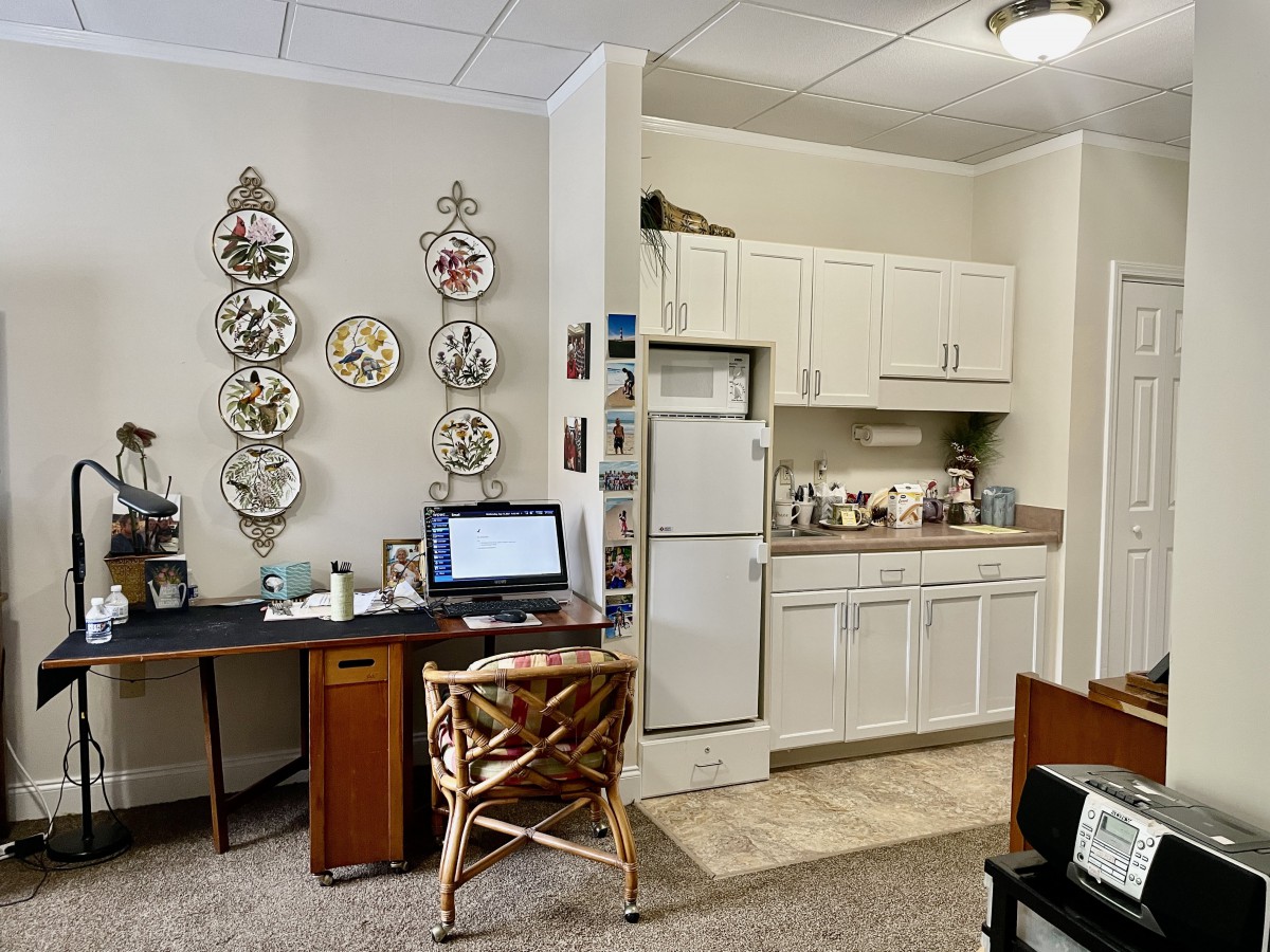A cozy office space with a desk and computer, connected to a small kitchenette adorned with decorative plates.