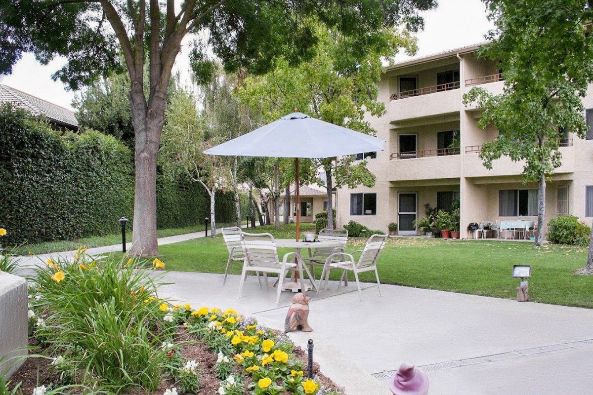 A serene outdoor patio area with a table, chairs, and umbrella set amidst a landscaped garden in front of an apartment building.