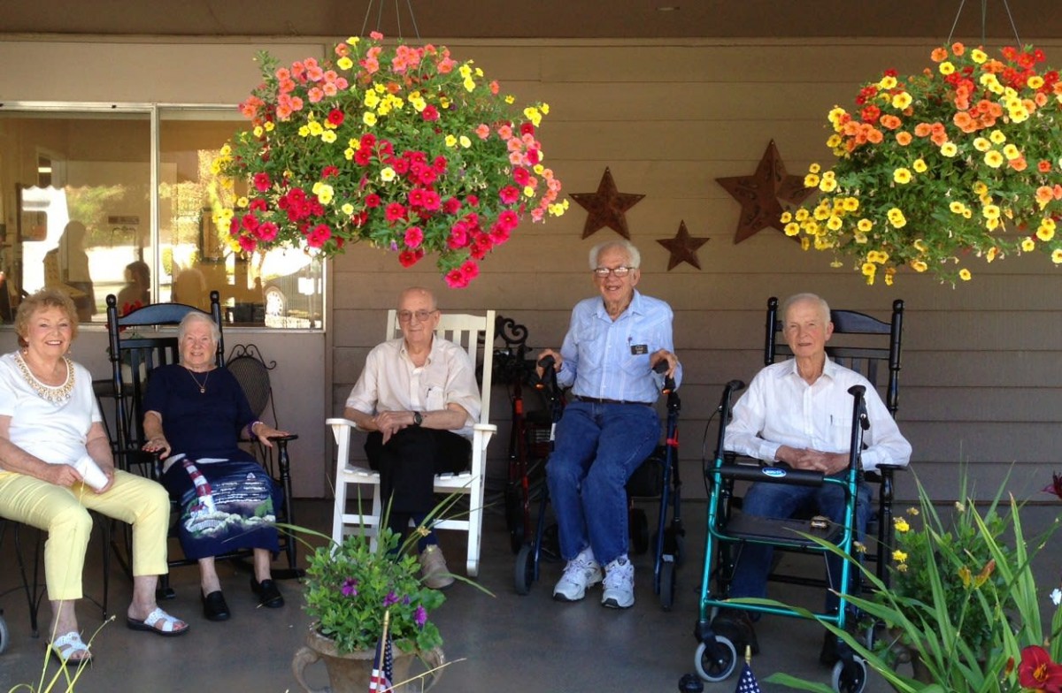 A group of elderly individuals sitting on a porch with hanging flower baskets and enjoying the outdoors.
