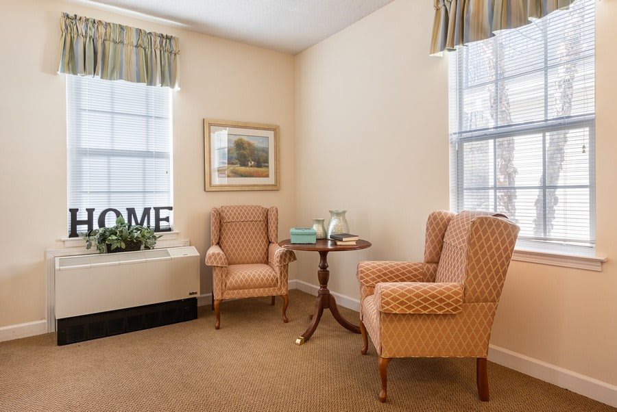 A cozy living room corner with two upholstered chairs, a small round table, and decor elements like a painting and window treatments.
