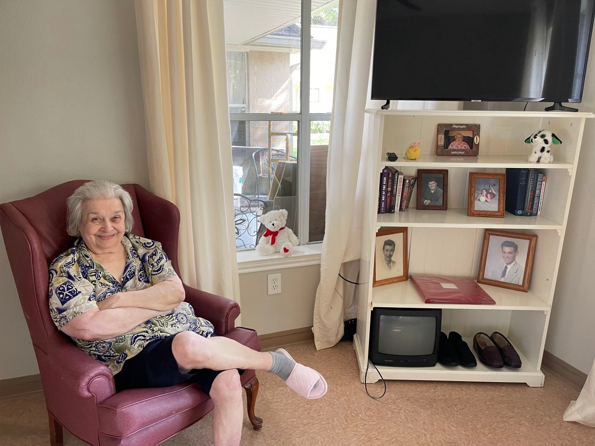 An elderly woman sitting comfortably in a living room next to a bookshelf with family photographs and a television.