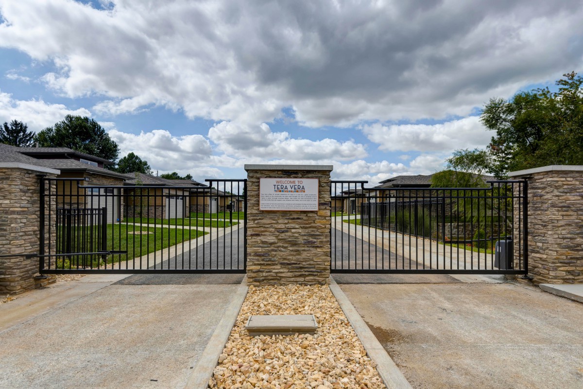 A gated entrance with a stone pillar under a cloudy sky, leading to a modern residential area.