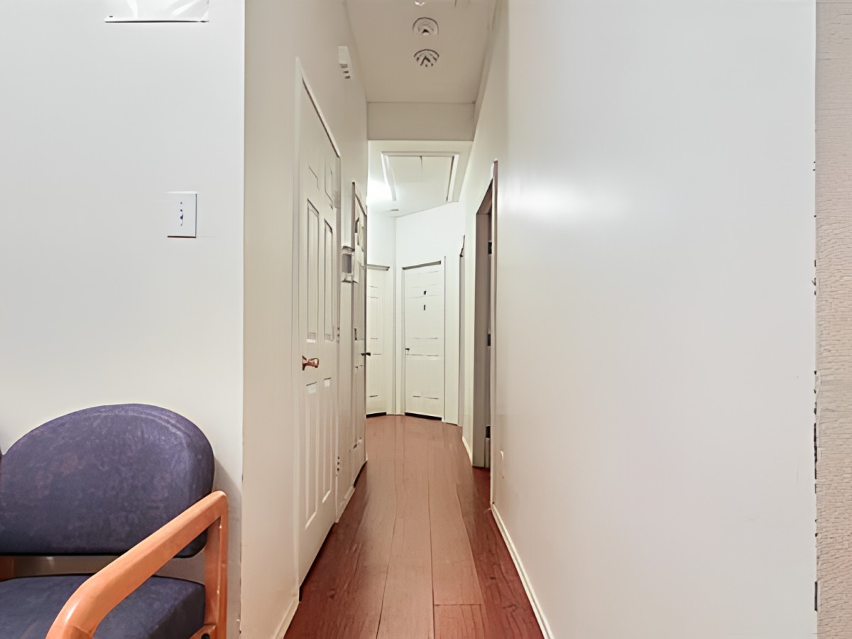 A narrow hallway with wooden flooring and white walls, featuring multiple doors and a chair on the left side.