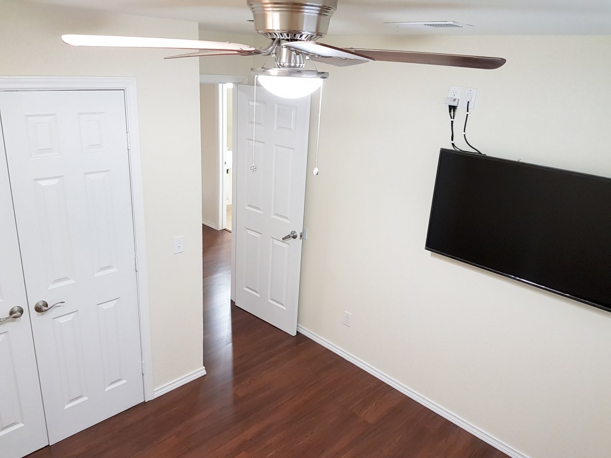 A room with a ceiling fan, wall-mounted TV, hardwood floors, and multiple doors.