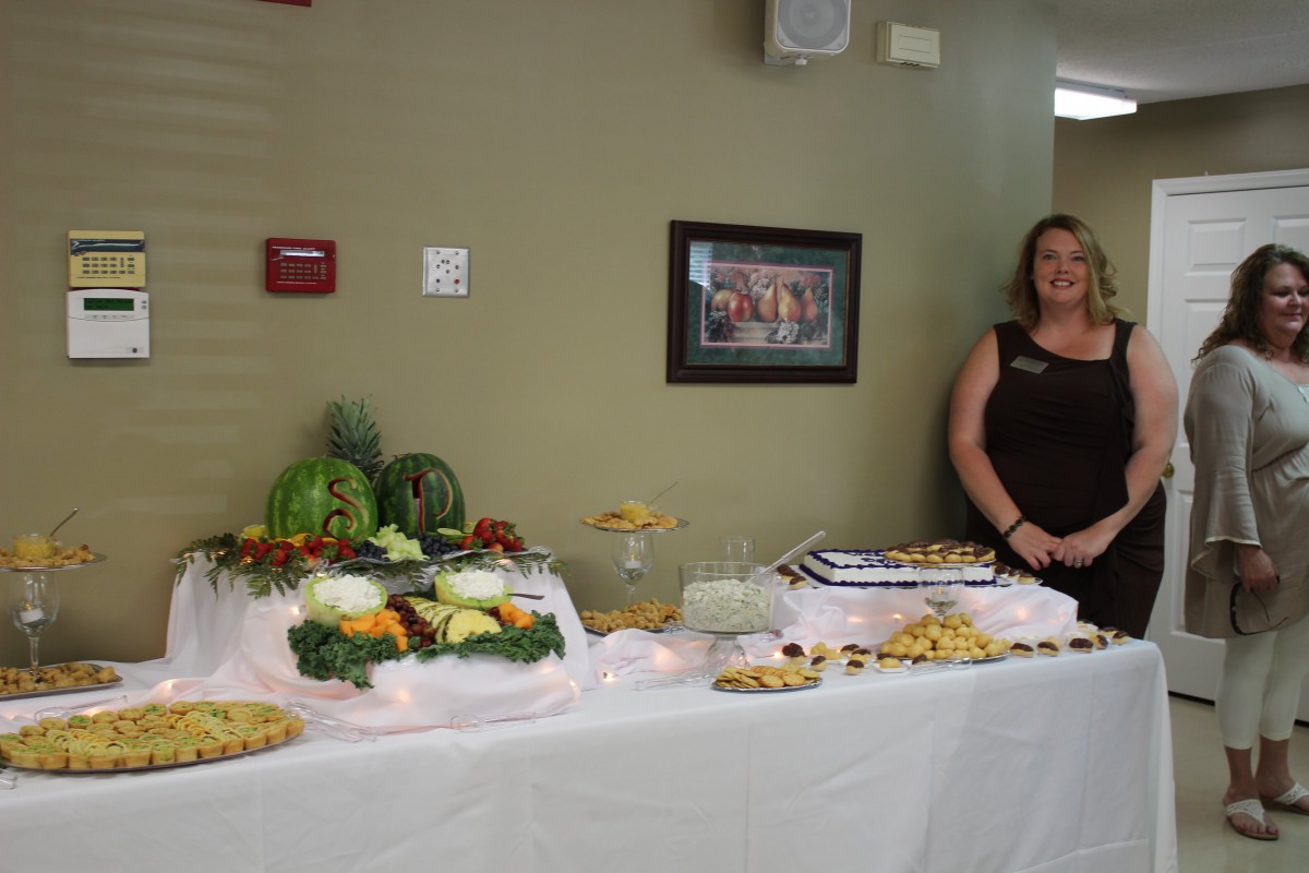A woman stands beside a buffet table with a display of fruits, vegetables, and various dishes.