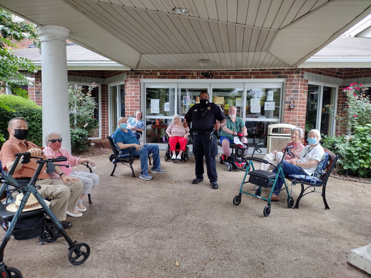 A police officer stands with a group of elderly individuals outside a nursing home, some seated in wheelchairs.