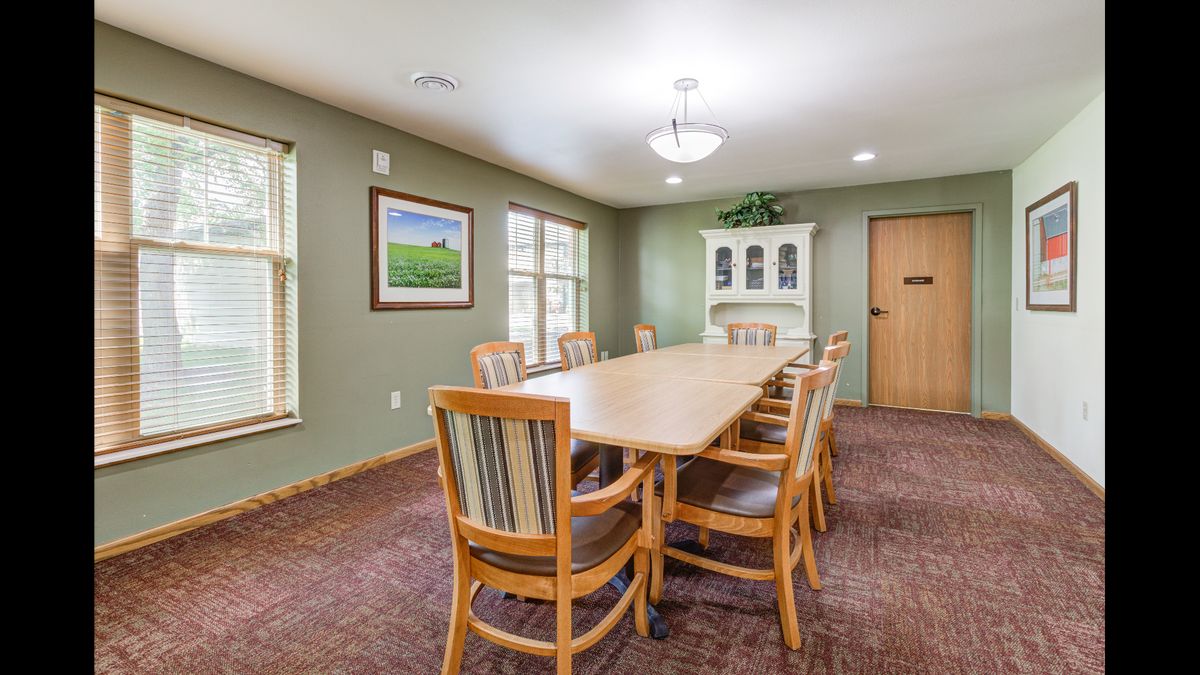 A well-lit conference room with a long wooden table surrounded by chairs, featuring a cabinet and windows.