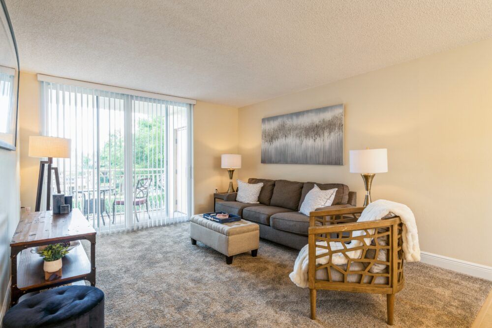 A cozy living room with a sofa, armchair, and coffee table, illuminated by natural light from a sliding glass door.