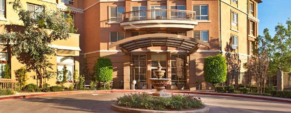 A modern building entrance with a glass canopy and a decorative fountain surrounded by plants.