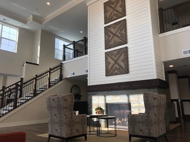 A modern living room interior with a high ceiling, featuring a staircase, artwork on the wall, and a cozy seating area by the fireplace.