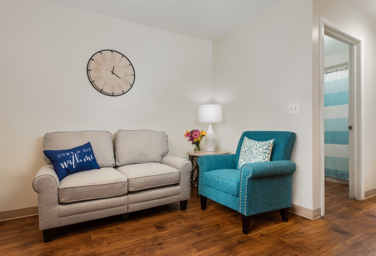 A cozy living room with a beige sofa and a blue armchair, accessorized with a wall clock and a side table lamp.