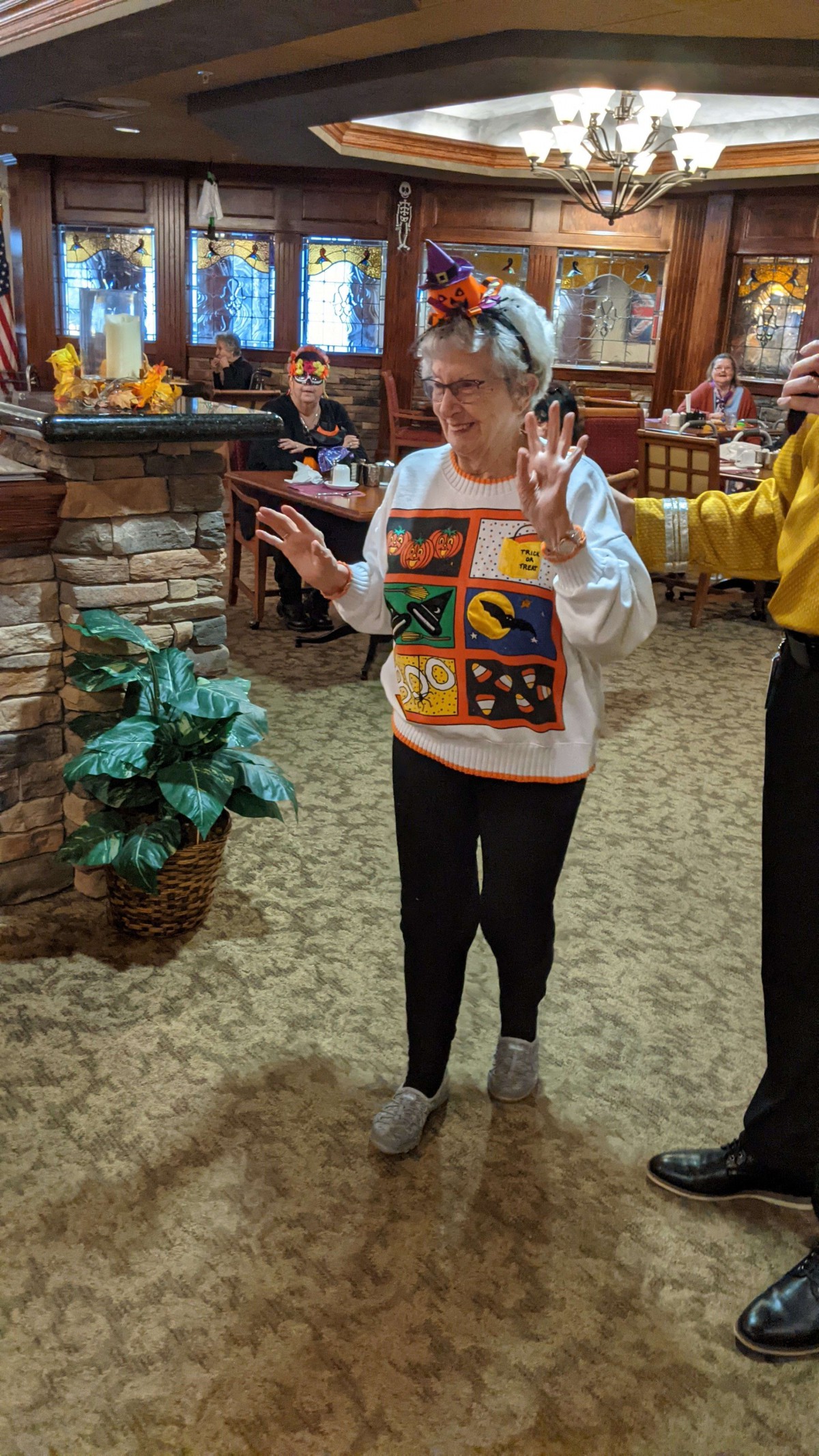 An elderly woman dances inside a restaurant, wearing a colorful sweater.