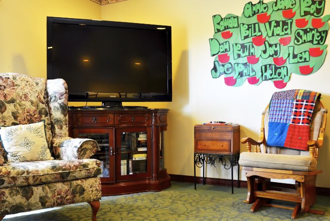 A cozy living room with a large television, floral-patterned armchair, and decorative wall art.