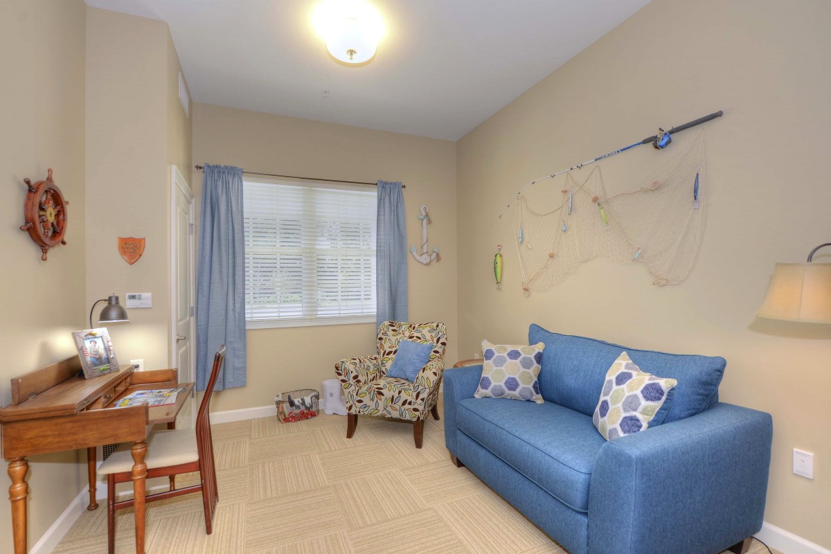 A cozy living room with a blue sofa, patterned armchair, and a small desk by the window.