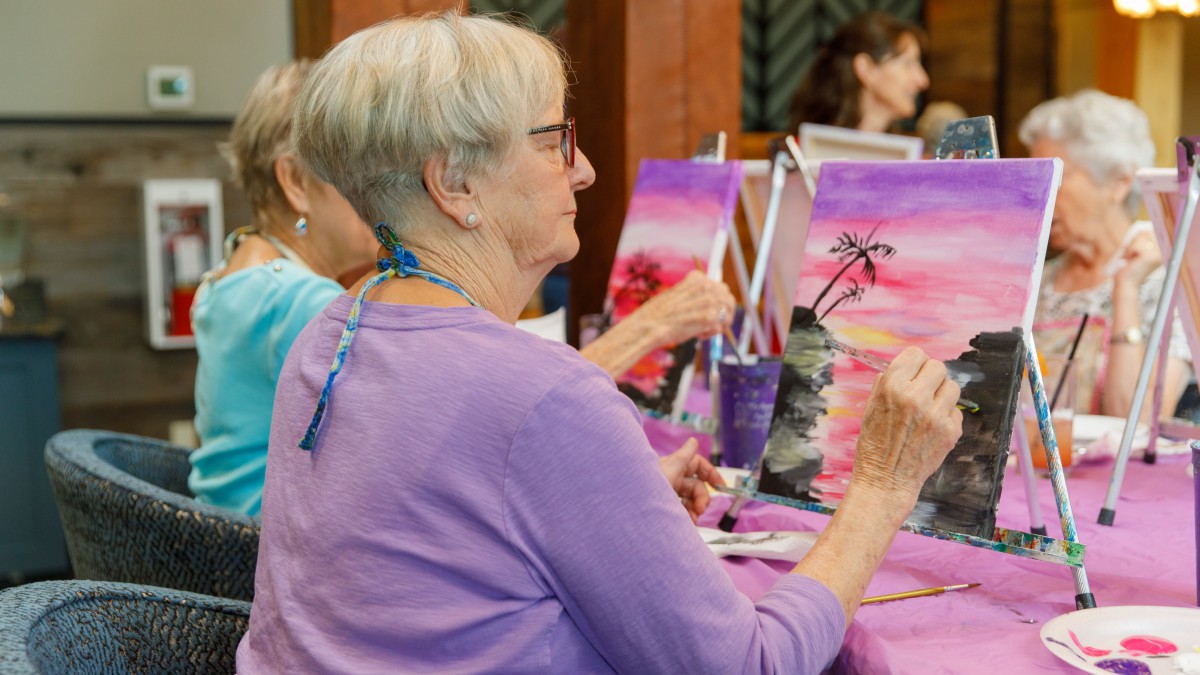 An elderly woman participates in an art class, painting on a canvas with vibrant colors.
