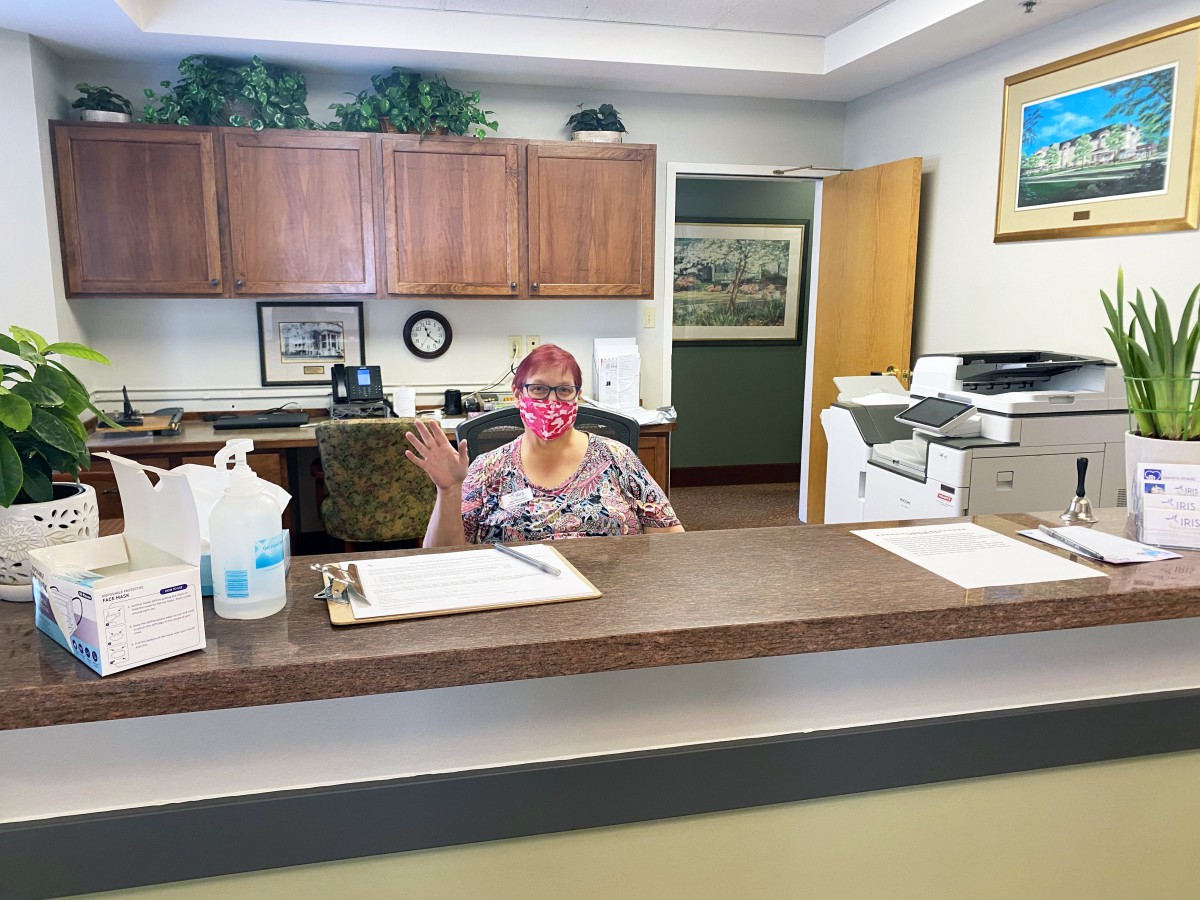 A receptionist wearing a mask sits at a desk in an office.