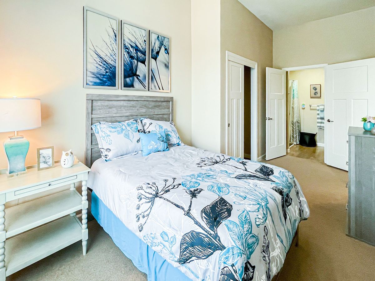 A cozy bedroom with a floral comforter and artwork above the bed, featuring a nightstand and a view into an adjoining room.