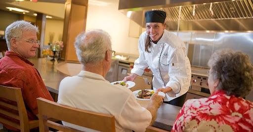 A chef serves a delicious meal to an elderly guest in a warm and welcoming kitchen environment.