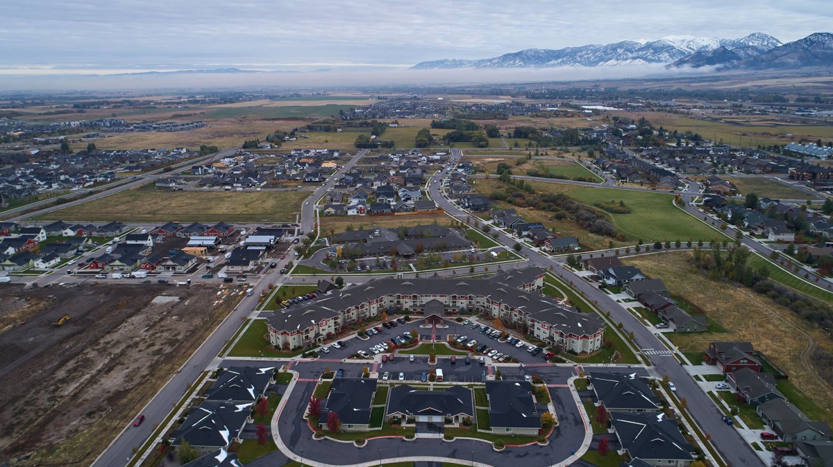 Aerial view of a suburban residential area with roads and houses, set against a backdrop of mountains under an overcast sky.