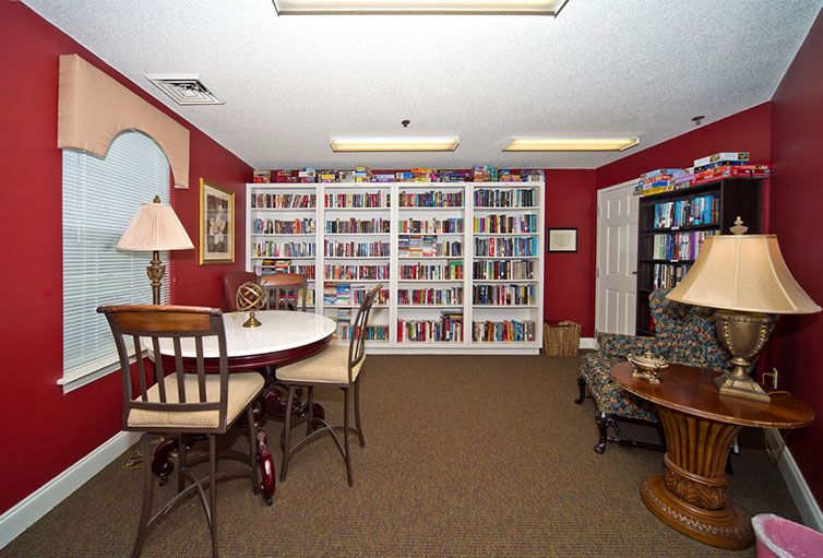 A cozy library room featuring bookshelves filled with books, a round table with chairs, and warm lighting.