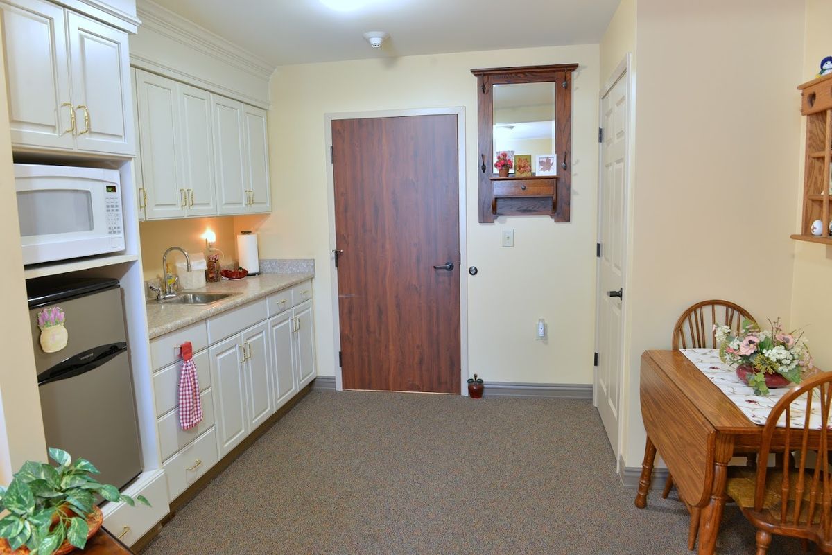 A cozy indoor kitchen with white cabinets, a sink, and a wooden door. The floor is covered with carpet.