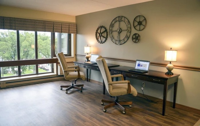 A modern office space with two desks and chairs, featuring wall decor and natural light from large windows.